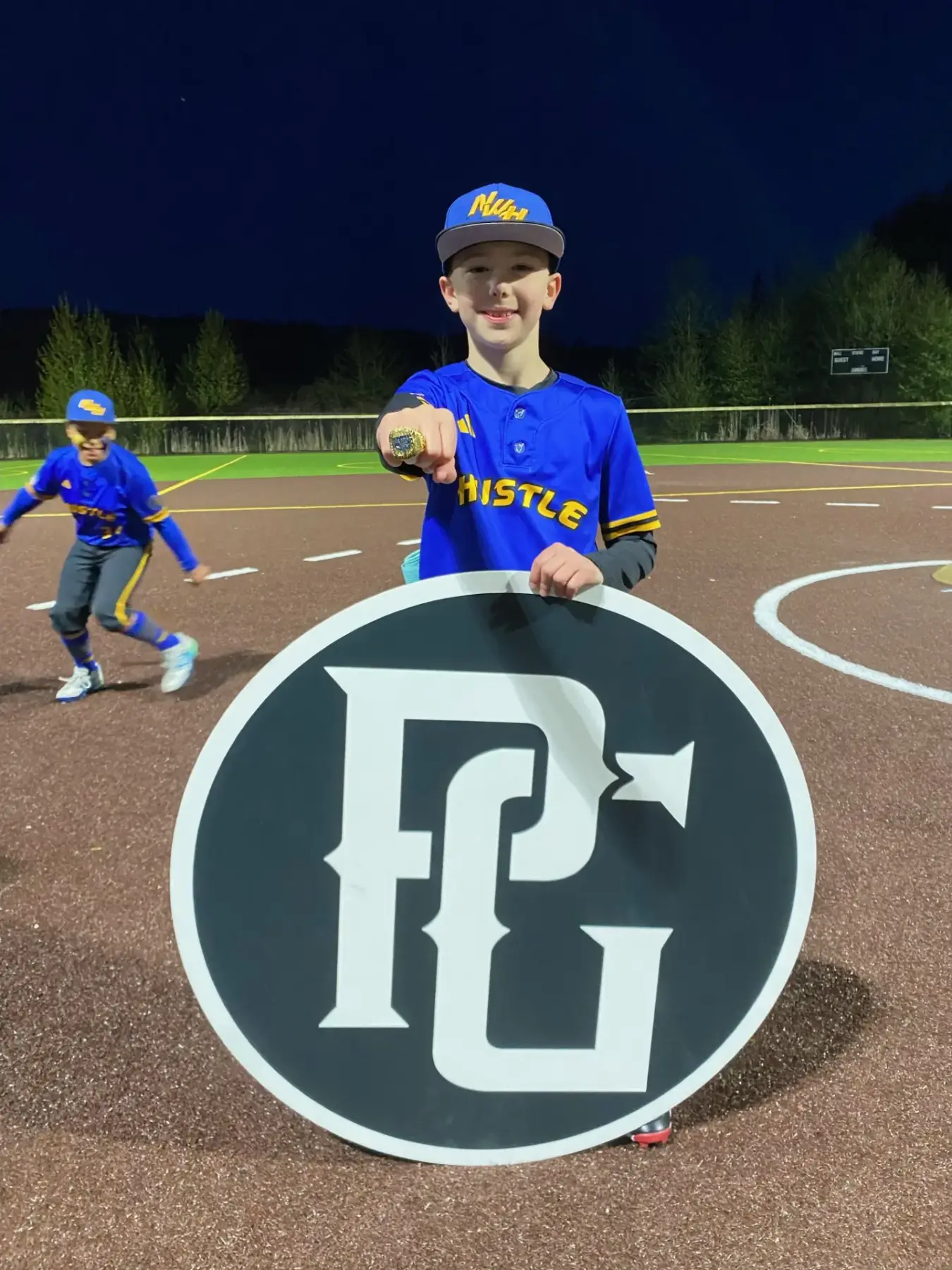Young player in blue uniform holding a large team logo sign and championship ring on the field at night.